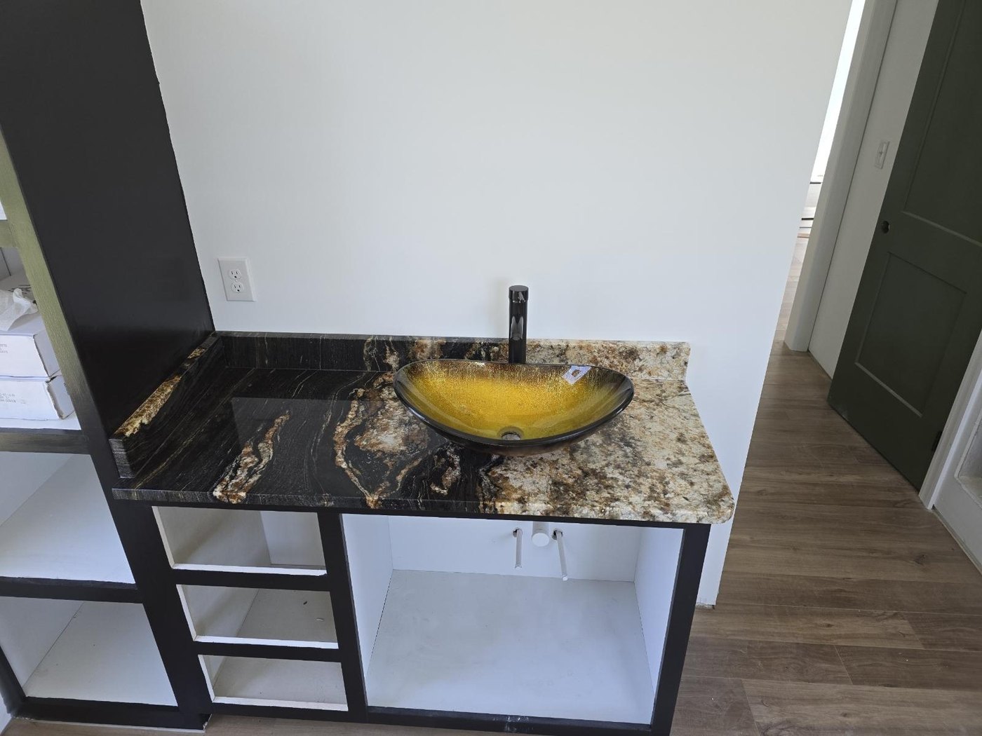 Vanity top in dark stone with gold and cream veining, a raised matching backsplash and an amber glass vessel sink with a black faucet, on a black-framed cabinet