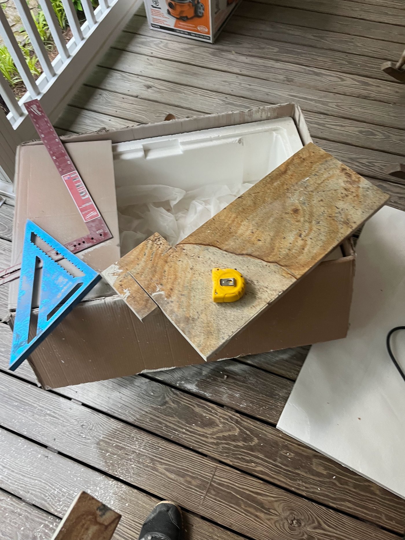 A stone sample piece resting in an open shipping box on a wood deck, beside a speed square, a folding ruler and a yellow tape measure