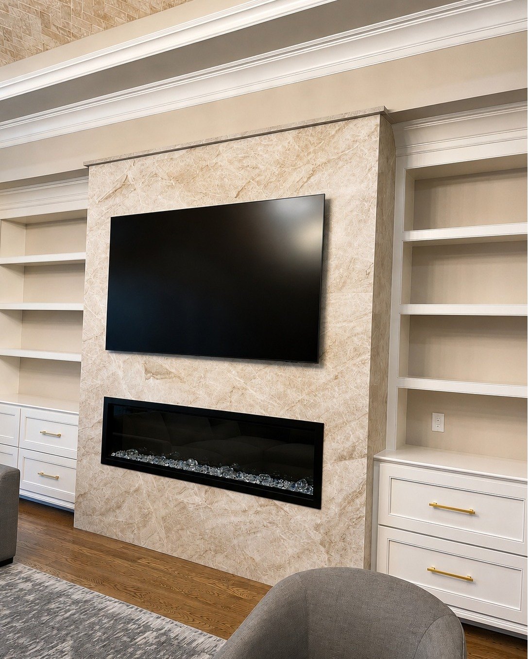 Living room feature wall faced in a cream-veined stone slab, with a wall-mounted television above a long linear fireplace, flanked by white built-in shelving and drawers