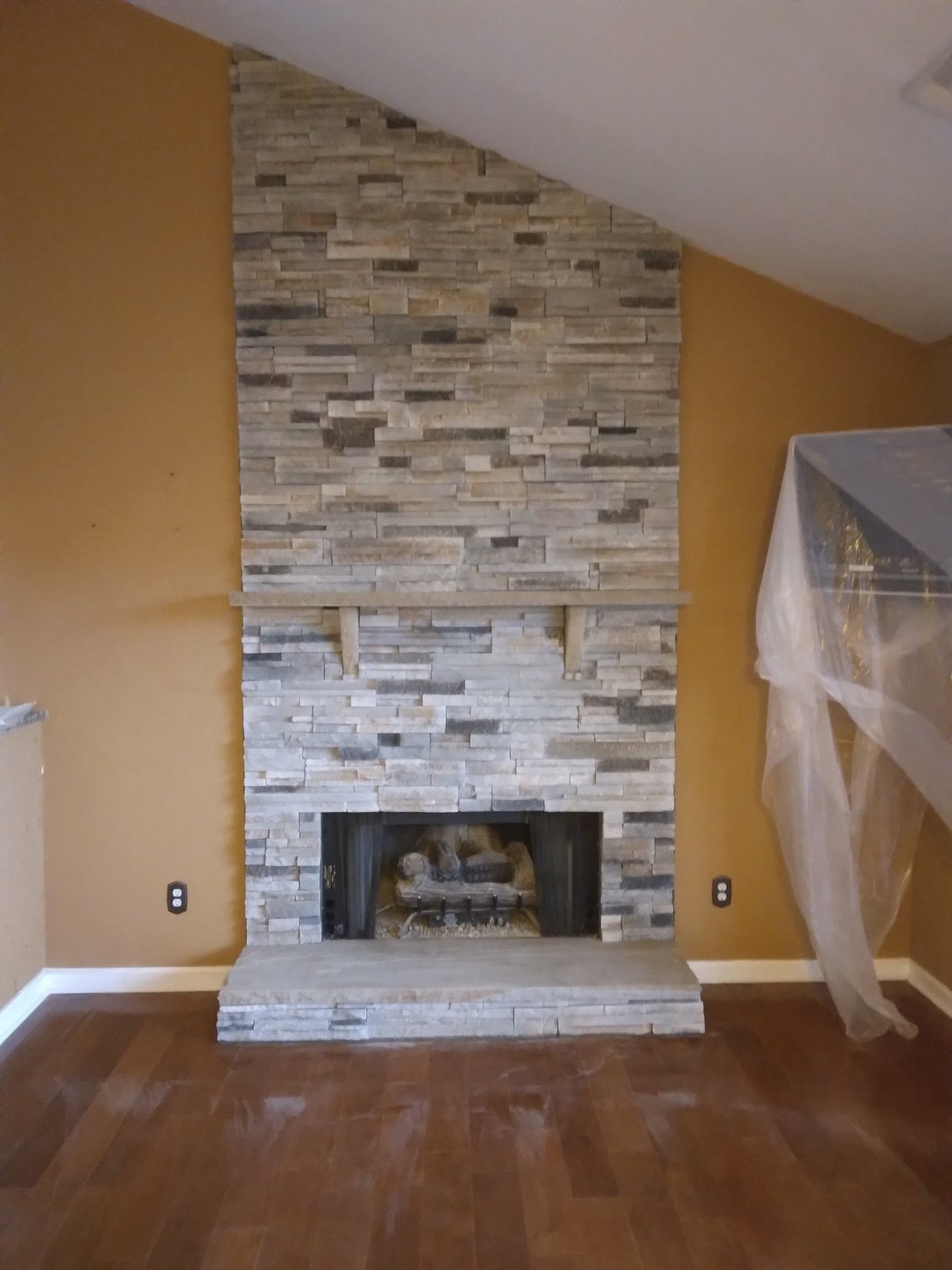 Floor-to-ceiling fireplace faced in stacked grey and tan ledgestone, with a stone mantel shelf on cut corbels and a raised stone hearth, in a room with a vaulted ceiling