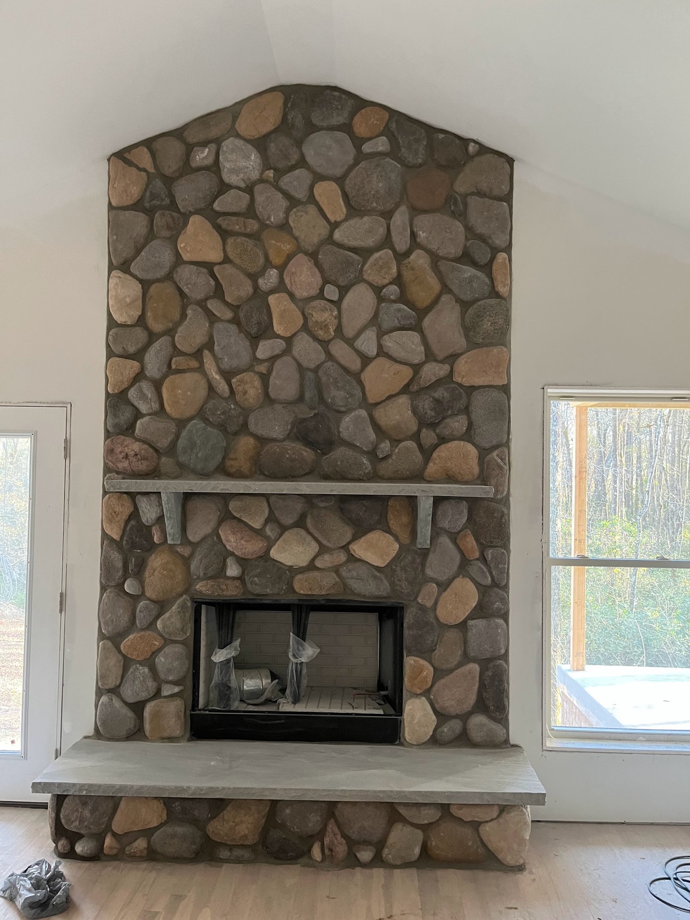 Rounded fieldstone fireplace running to a gabled top against a vaulted ceiling, with a cast mantel shelf and a full-width cast hearth, seen from across the room