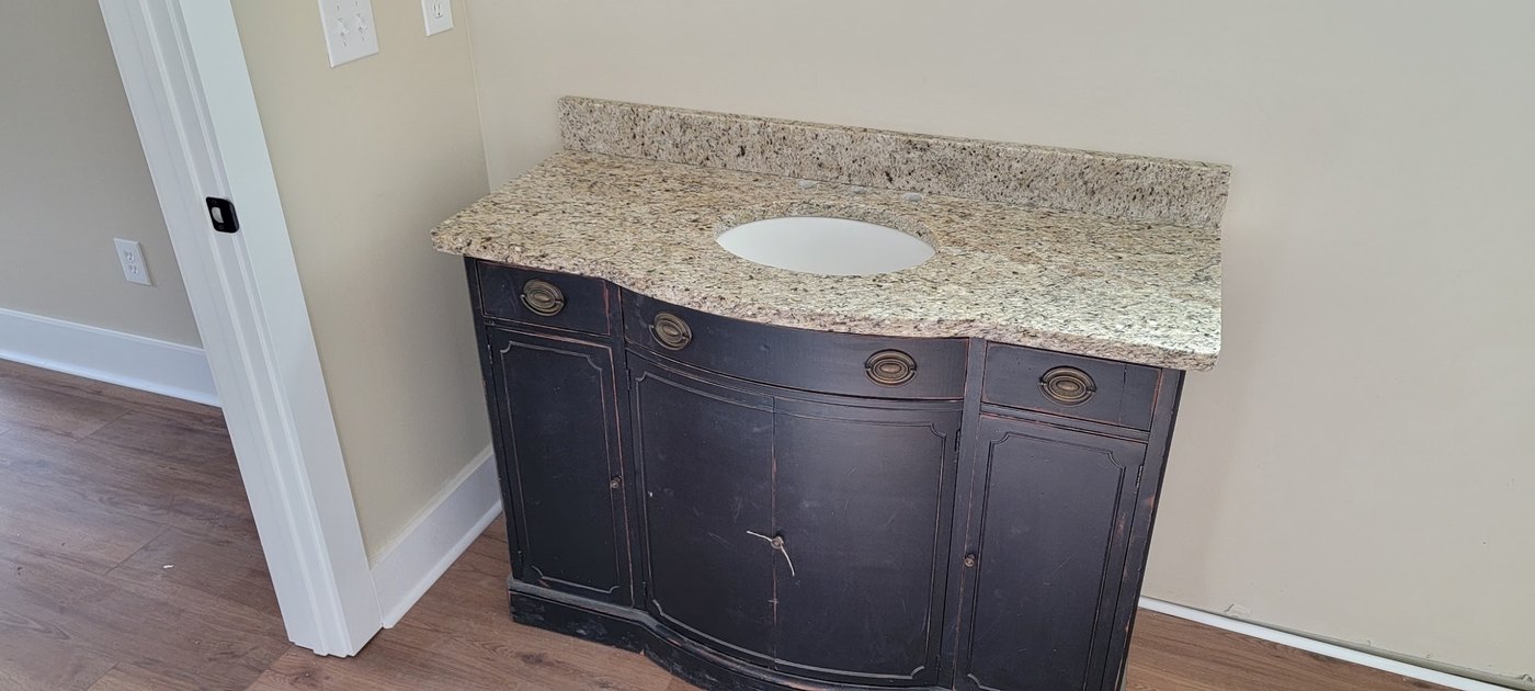 Antique black sideboard rebuilt as a bathroom vanity, topped with a speckled cream stone counter cut for an oval undermount basin, in an empty room