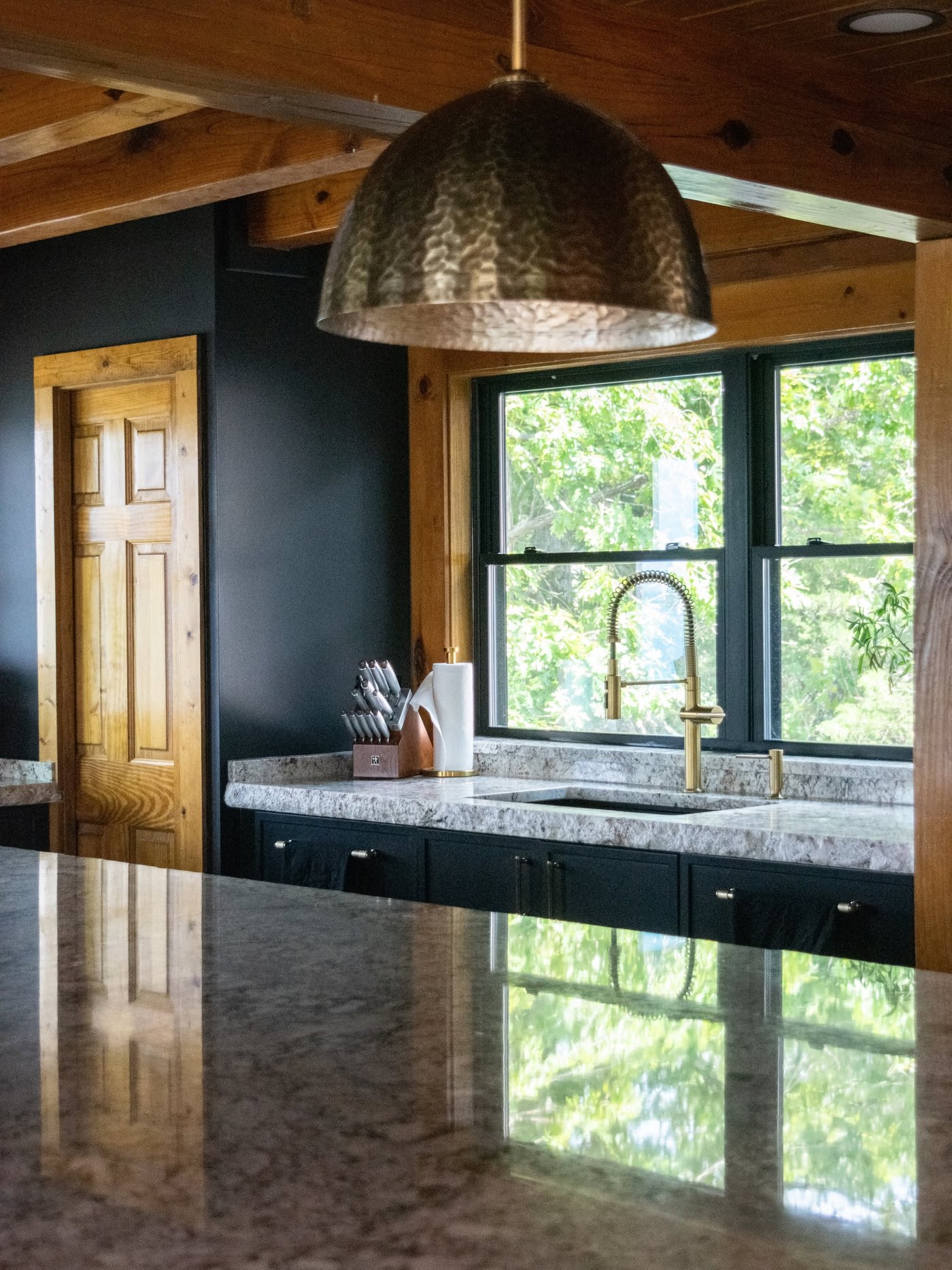 Log home sink run in veined granite under a window, with a brass faucet, dark cabinetry and a hammered pendant above