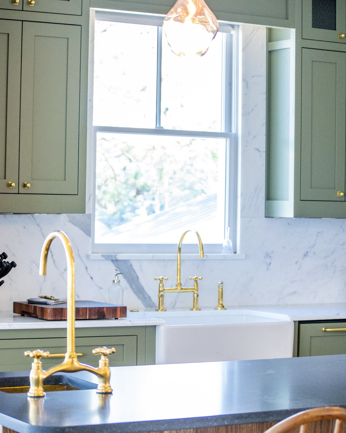 Green kitchen with a white apron-front sink, brass bridge faucet and a white veined stone backsplash running up behind the window