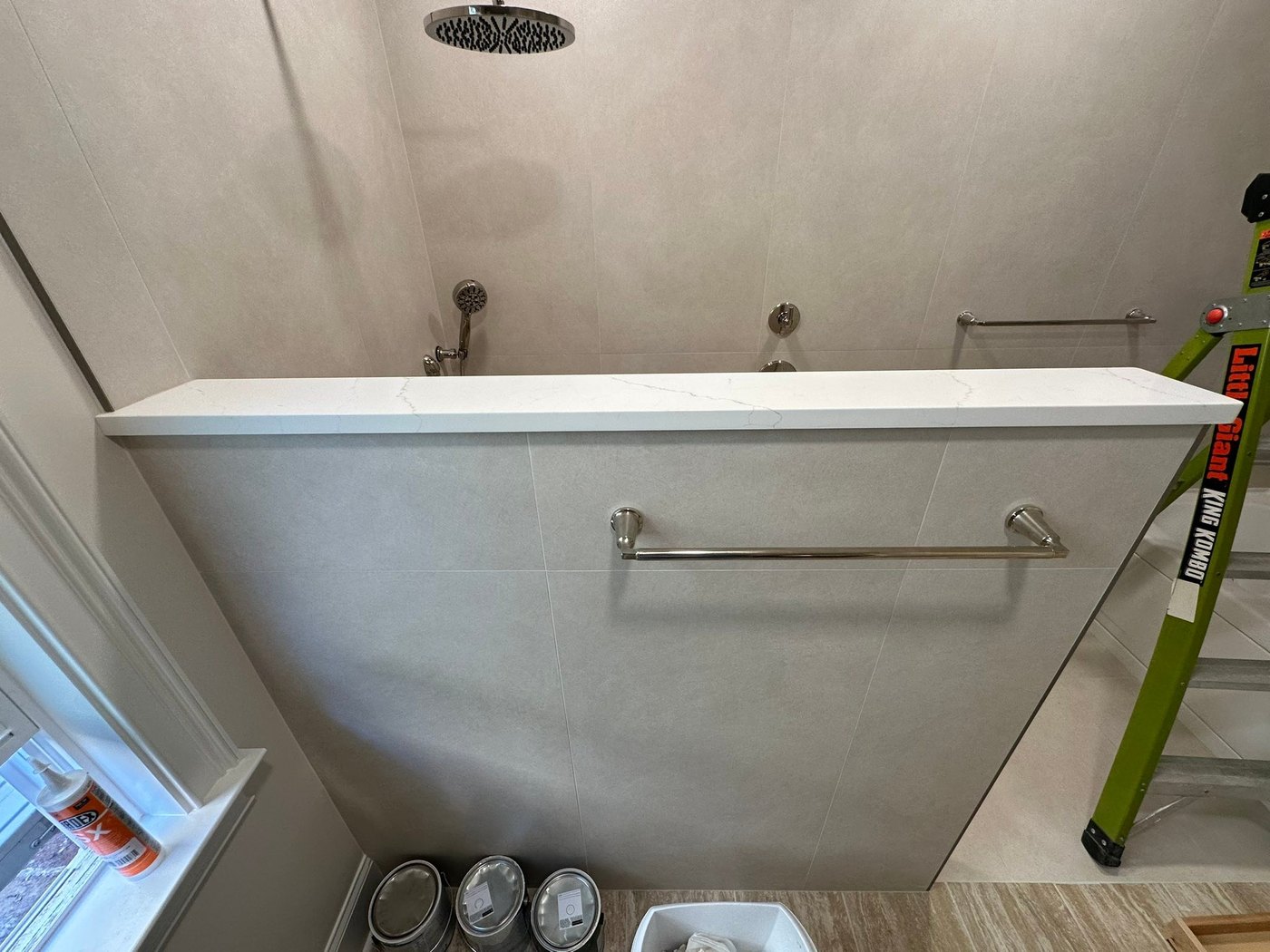 Walk-in shower with a white stone cap on the half-wall, a rain shower head and tiled walls, still under construction