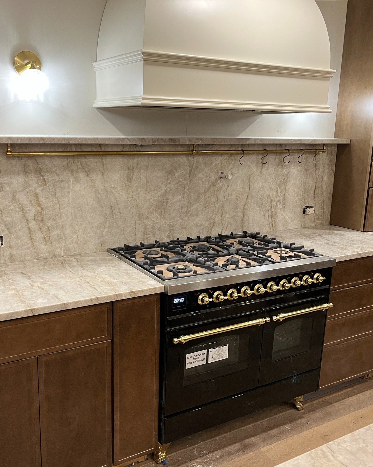 Full-height stone slab backsplash with soft brown veining behind a black and brass range, under a cream plaster hood, with walnut cabinetry