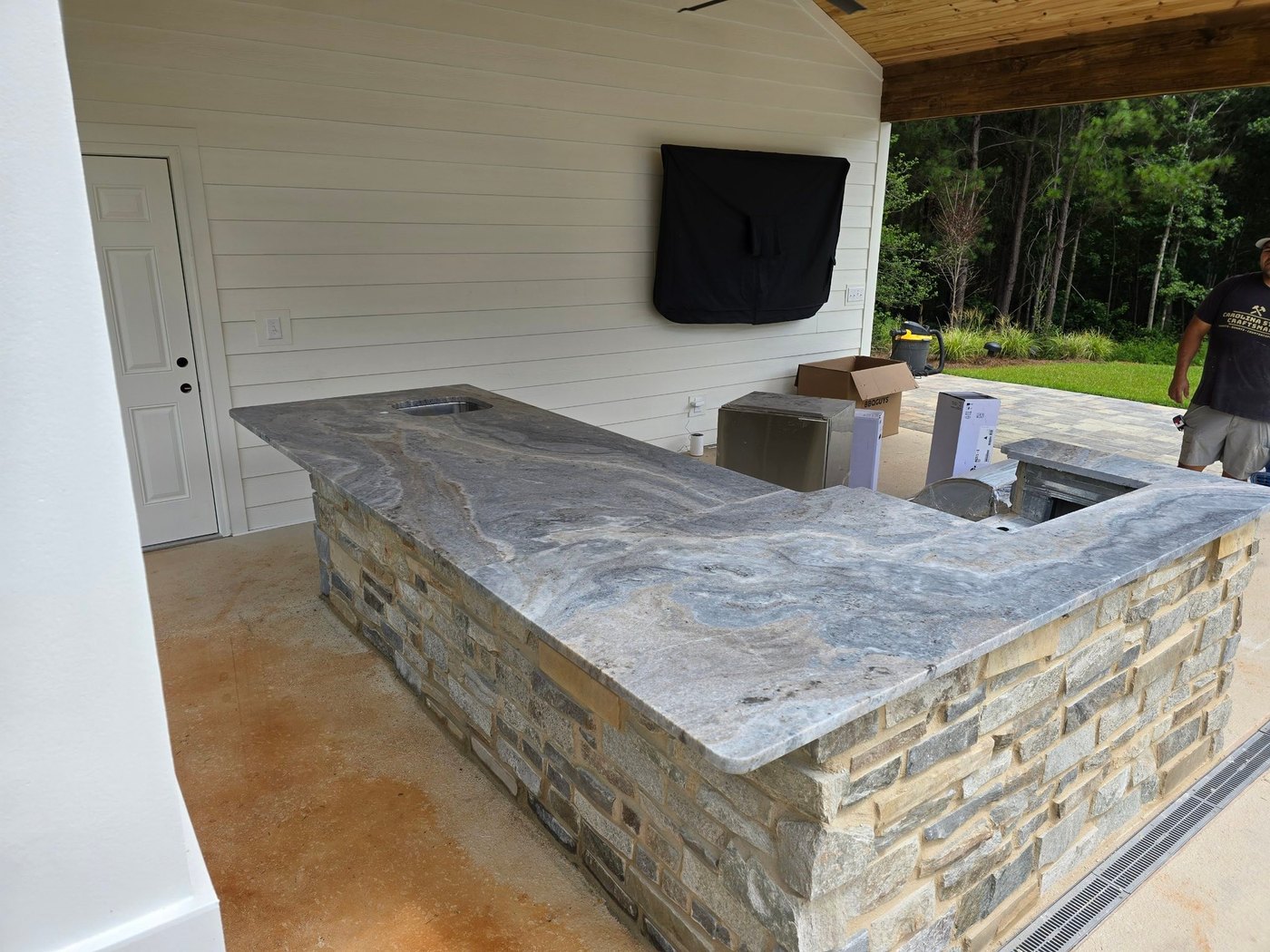 Covered outdoor bar with a stacked-stone base and a grey and tan veined stone top, an outdoor TV on the shiplap wall and pines beyond