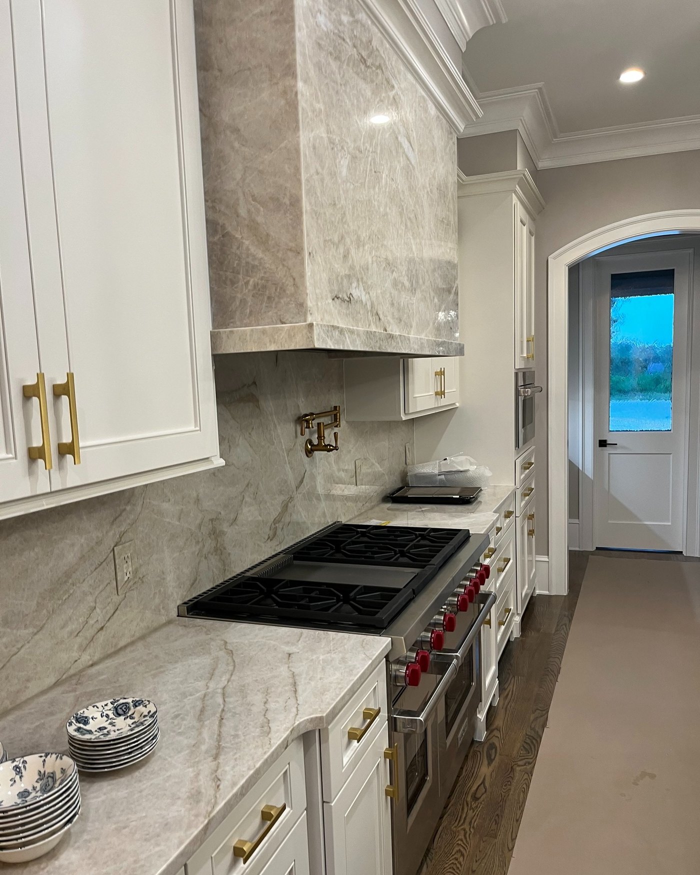 White kitchen with a stone-clad range hood and a matching veined slab backsplash behind a gas cooktop, brass hardware