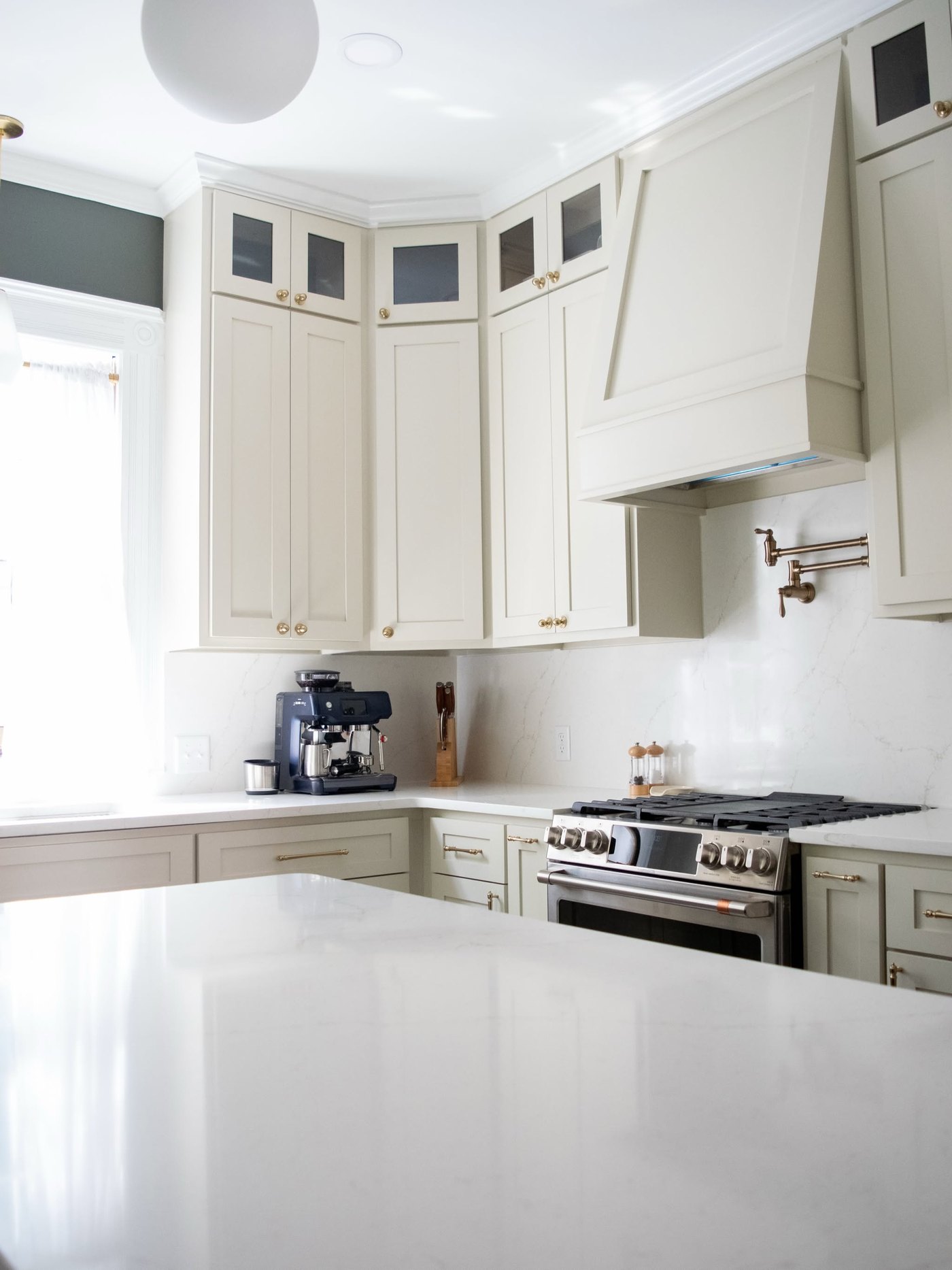 Cream kitchen with a painted hood, glass-front upper cabinets and a plain white counter running past a stainless range