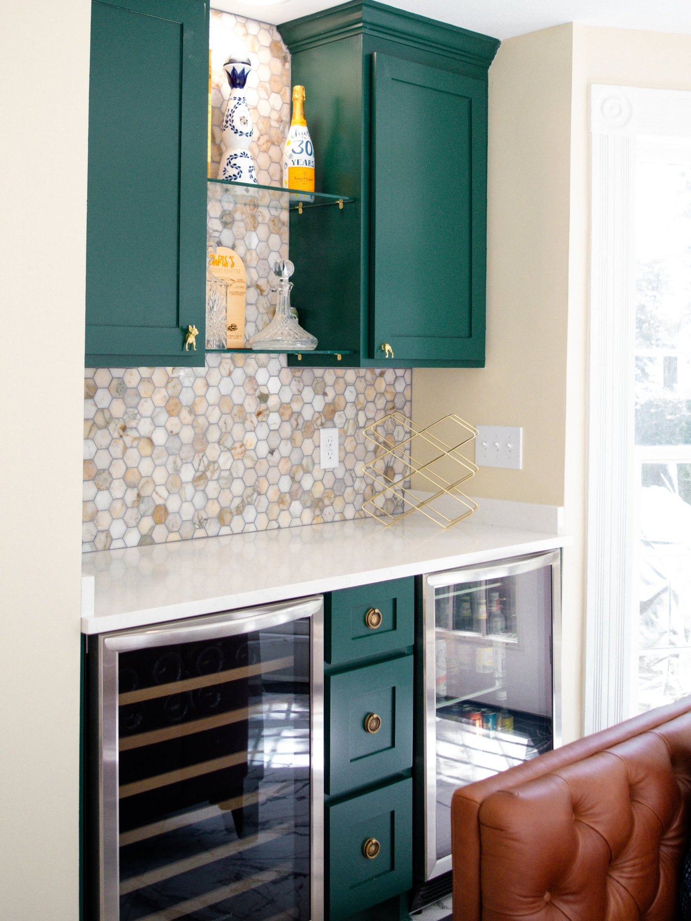 Beverage bar with deep green cabinets, a hexagonal stone mosaic backsplash, a white counter and two glass-front wine coolers
