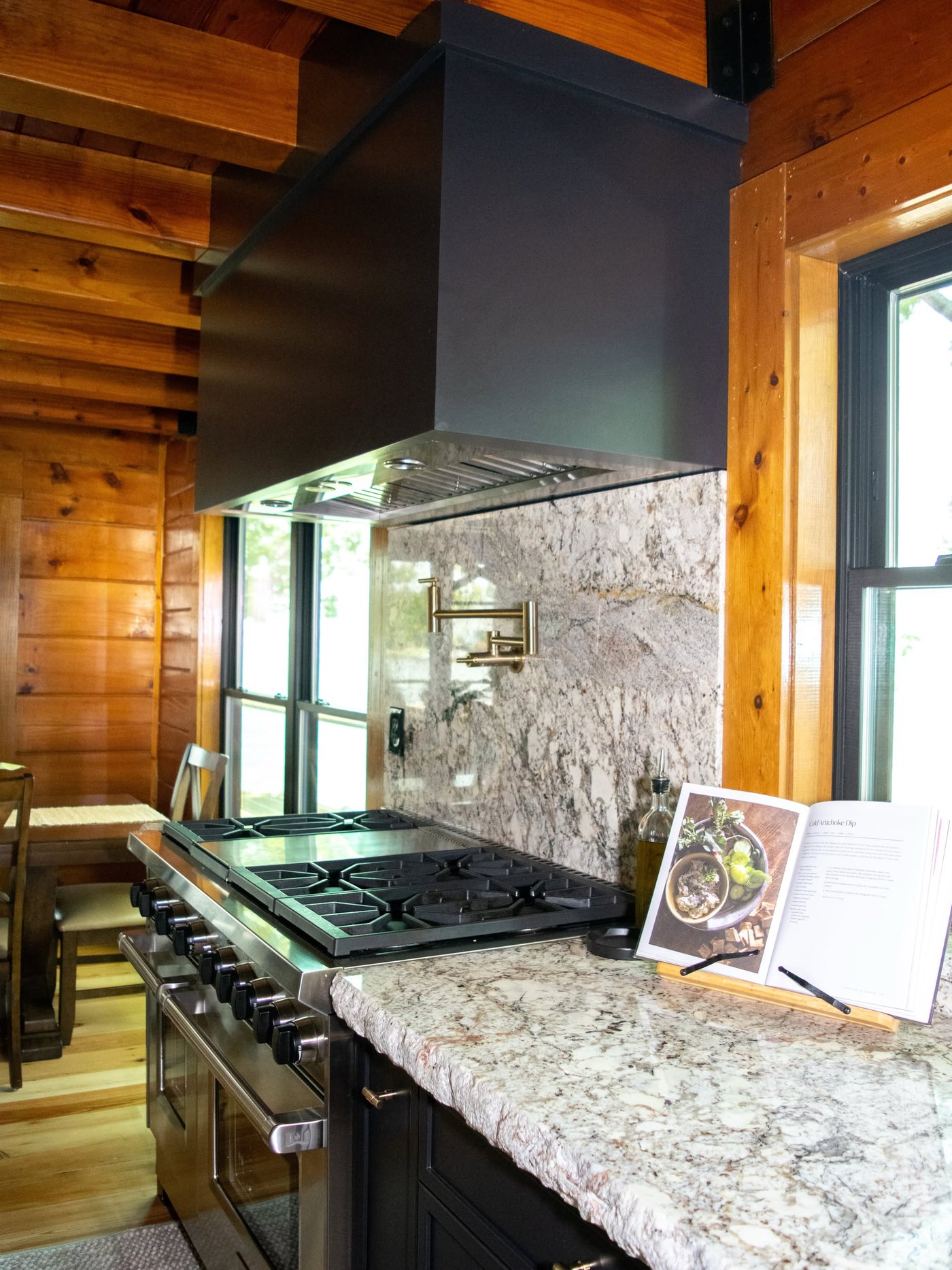Log home kitchen with a veined granite counter and matching slab backsplash behind a stainless range, under a steel hood and timber beams