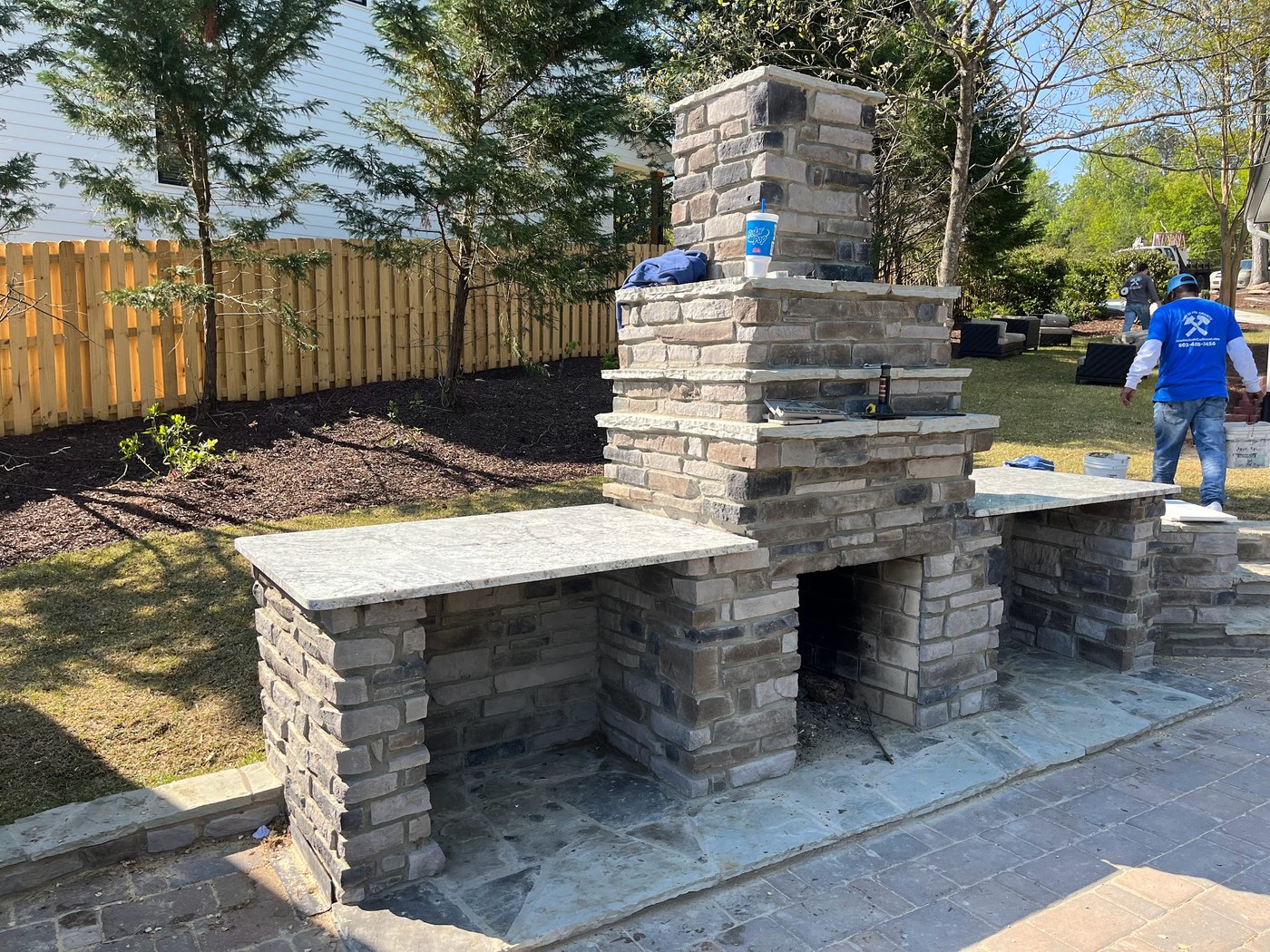 Outdoor stone fireplace under construction with a tall chimney and stone bar counters running out to either side, over a paver patio