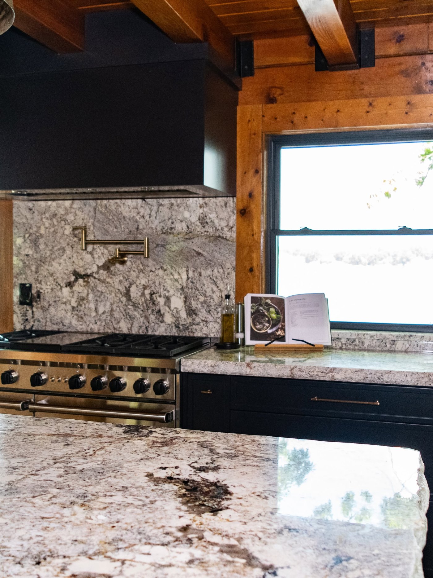 Wider view of the same log home kitchen: granite island in the foreground, range wall and slab backsplash beyond, cedar walls
