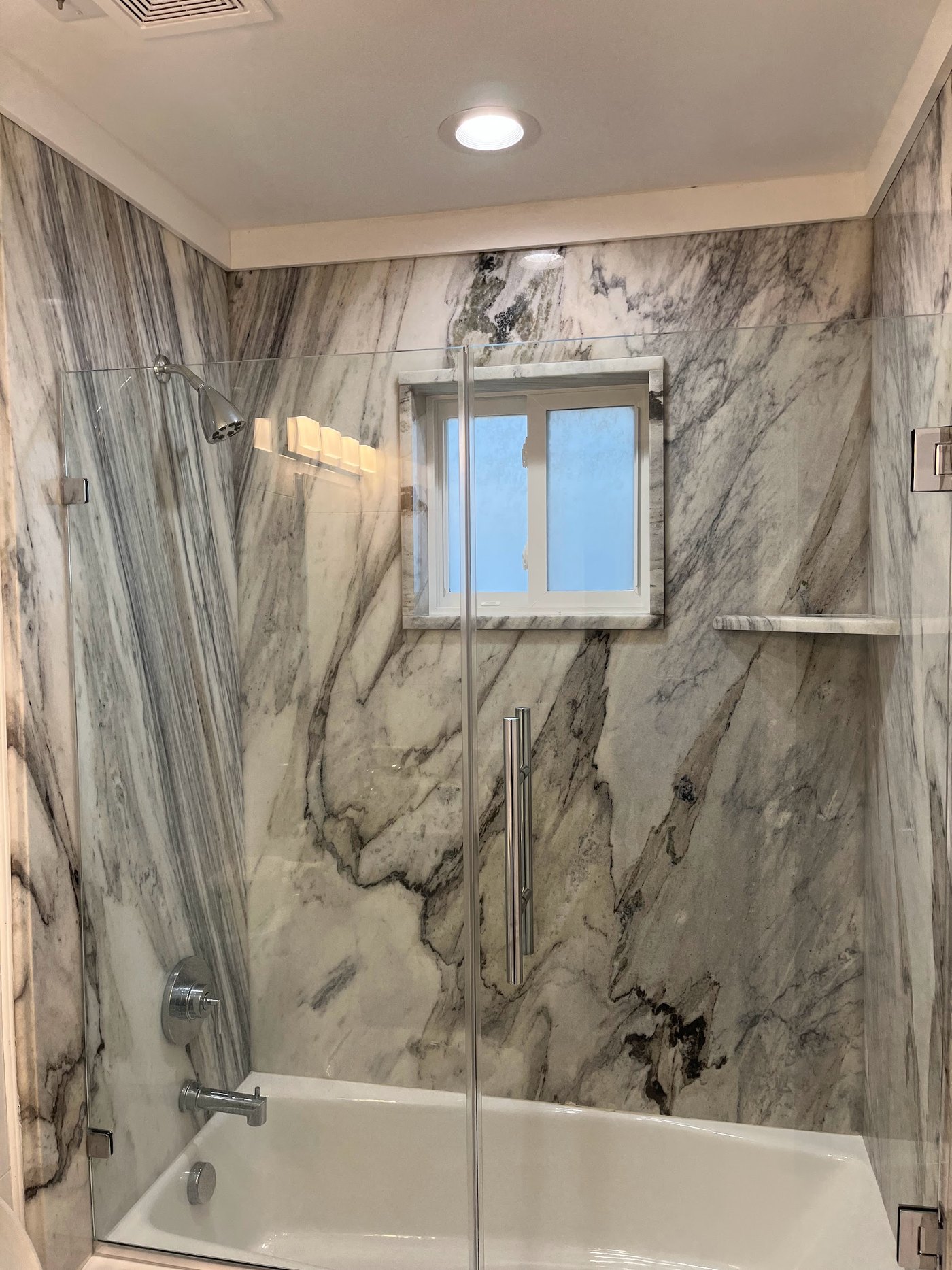 Tub and shower surround clad in grey-veined marble slab, continuous across the walls, behind a glass door