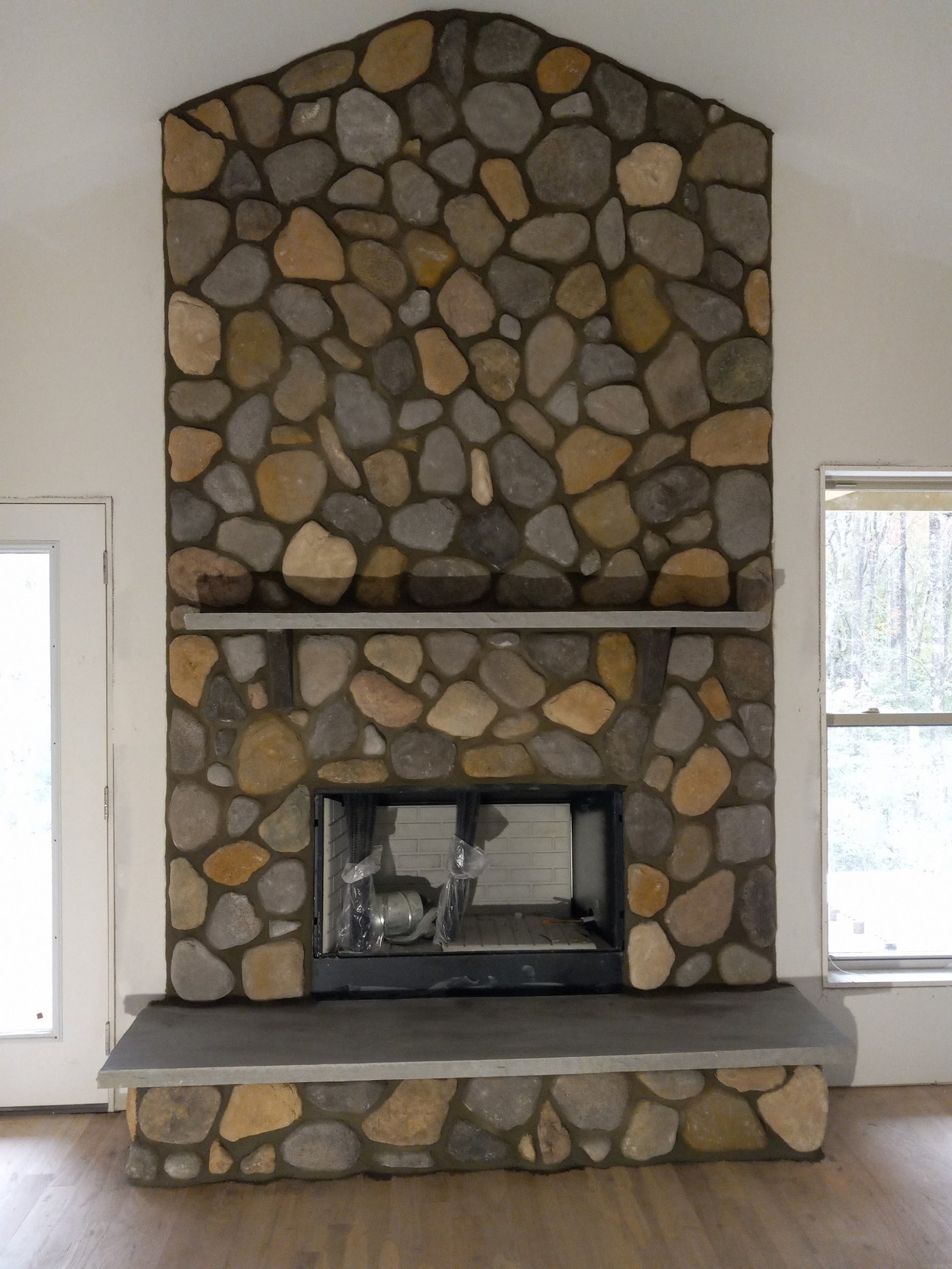 Floor-to-ceiling rounded fieldstone fireplace with a gabled top, a cast hearth and a matching mantel shelf, built into an interior wall