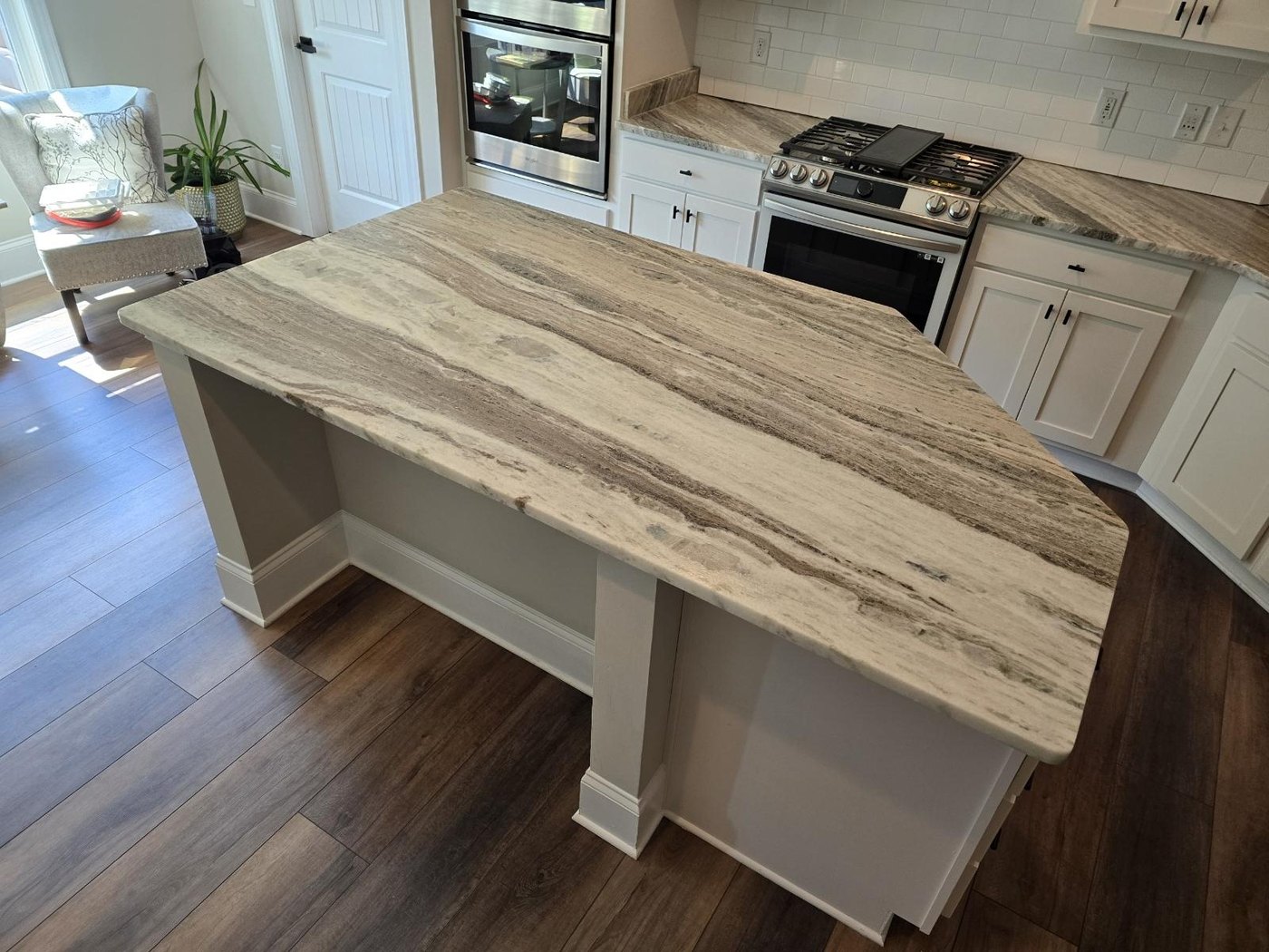 Kitchen island topped with a single grey-and-cream veined stone slab with a thick eased edge and a clipped corner, in a white kitchen with a gas range and matching perimeter counters