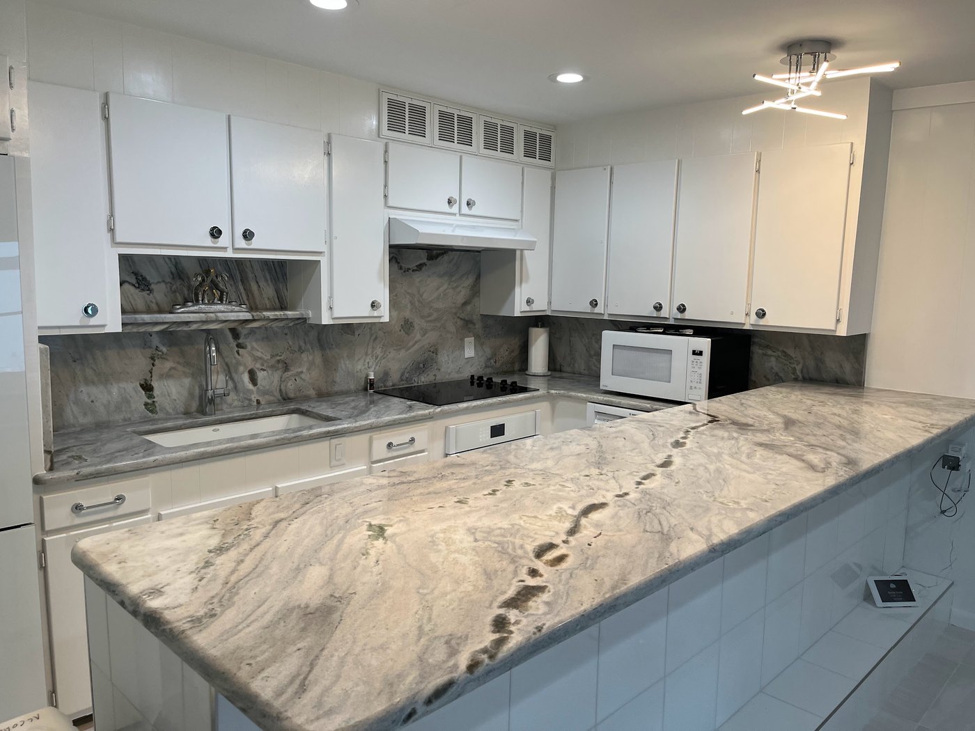 White kitchen with grey and brown veined stone counters wrapping a peninsula, a matching full-height slab backsplash behind the range and an undermount sink