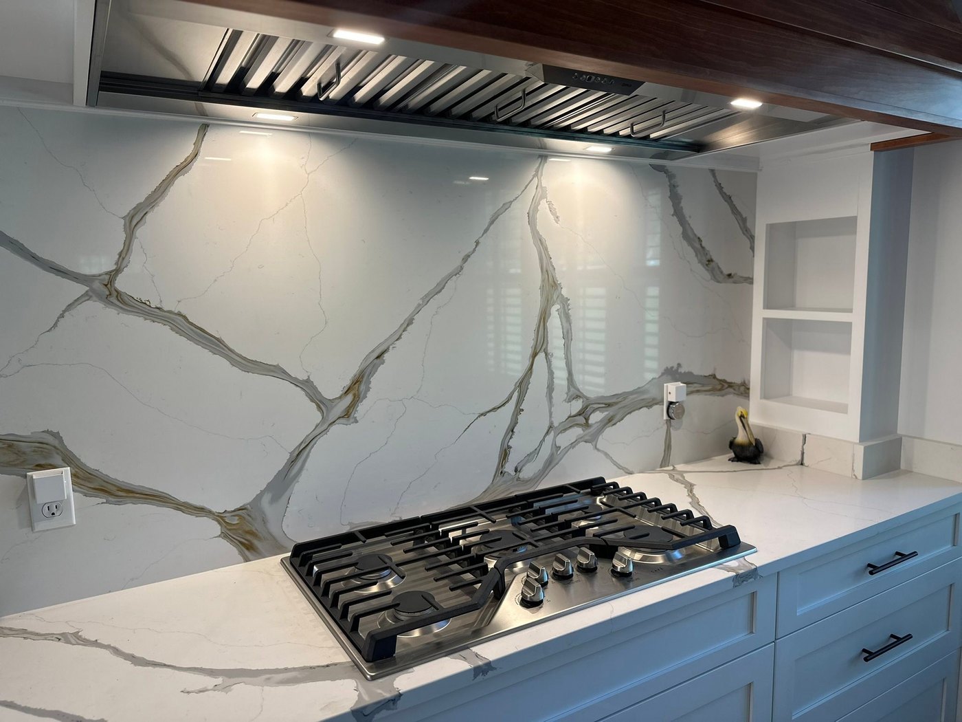 Full-height white stone slab backsplash with dramatic grey and gold veining behind a gas cooktop and a stainless hood, over pale blue cabinetry