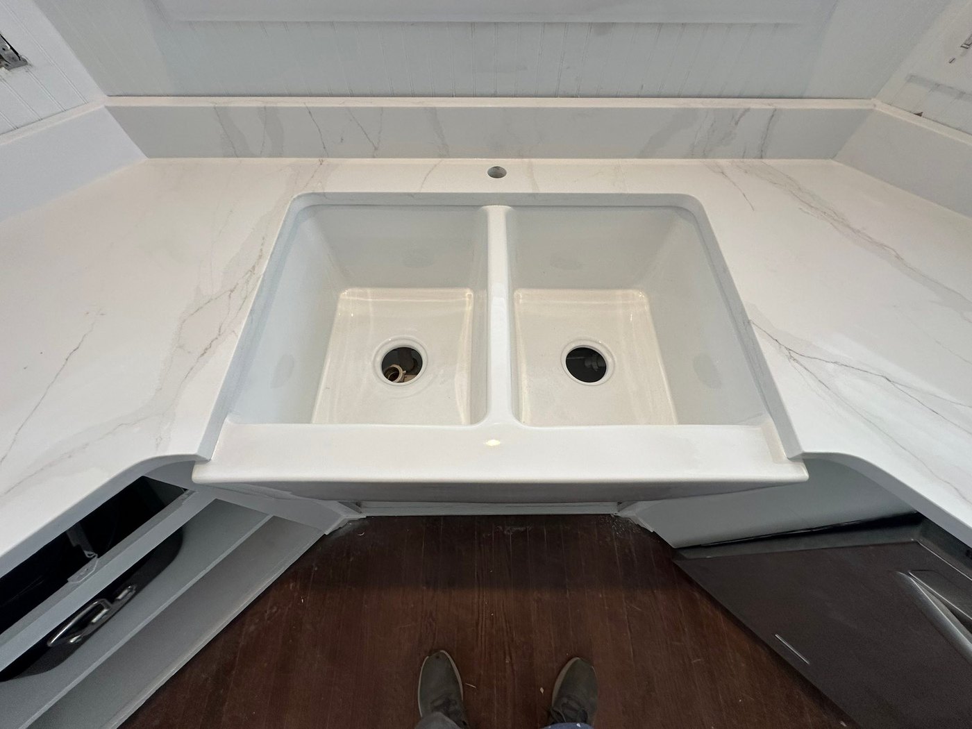 White apron-front double farmhouse sink set into a white veined stone counter with a matching slab backsplash, above a dark wood floor