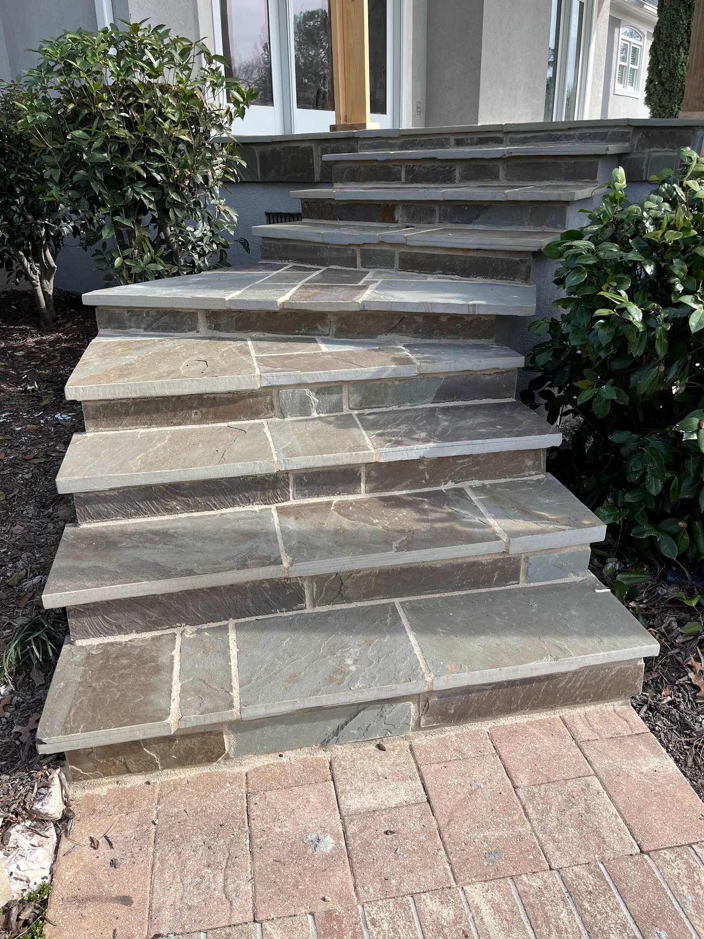 Wide flagstone steps with cut stone risers climbing to a house entry, meeting a brick paver walkway at the bottom