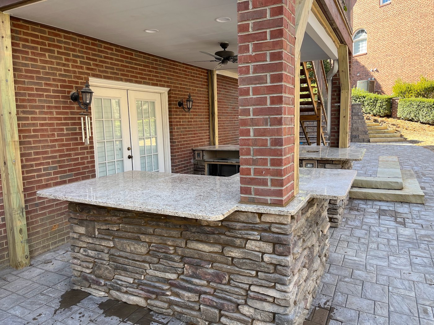 Covered brick patio with an L-shaped outdoor bar, its base faced in stacked stone and topped with a pale speckled stone counter, over a stamped concrete floor