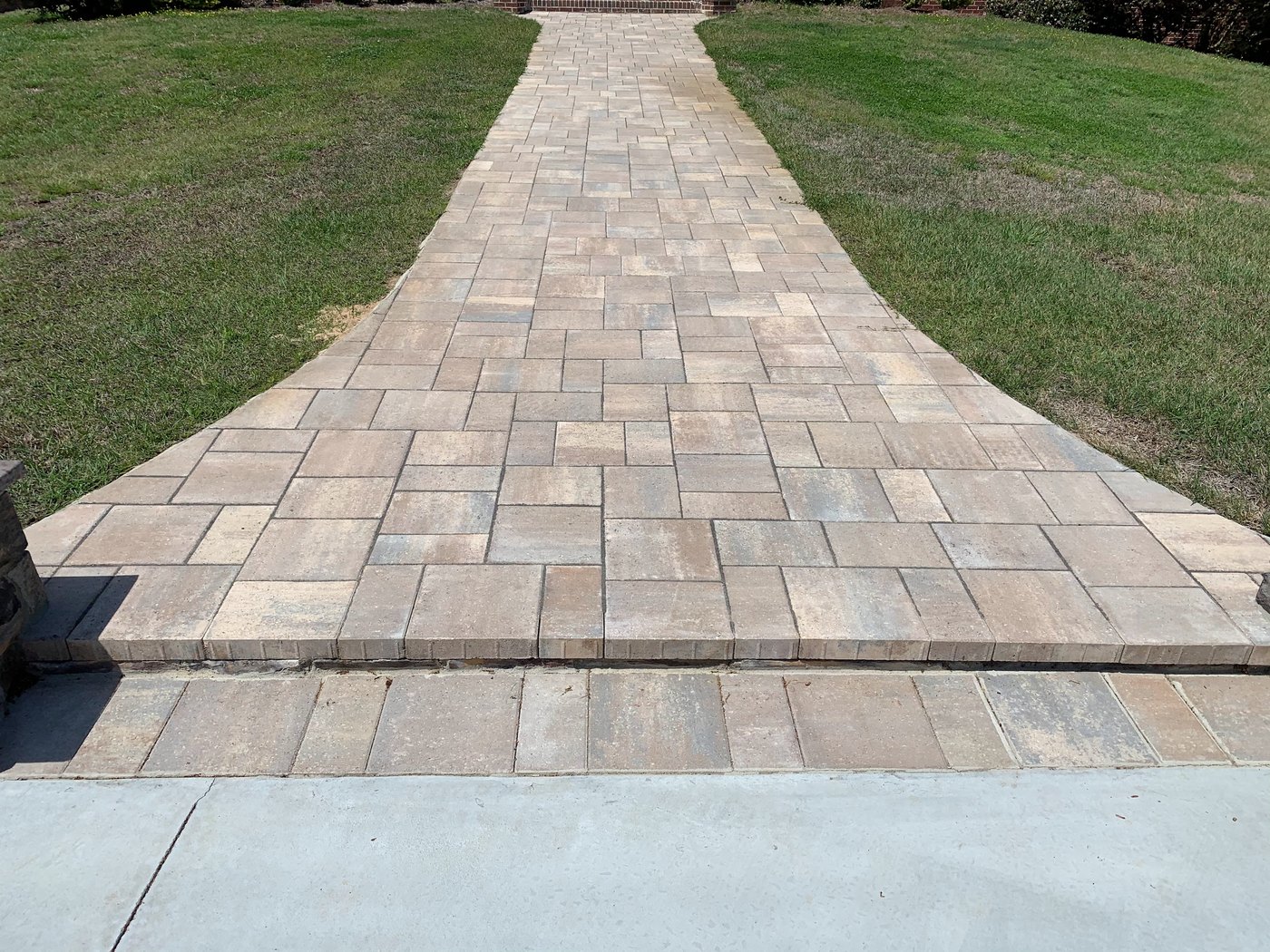 Close view of a tan and grey paver walkway in a running bond pattern with a soldier-course border, meeting a concrete driveway at a stone wall corner