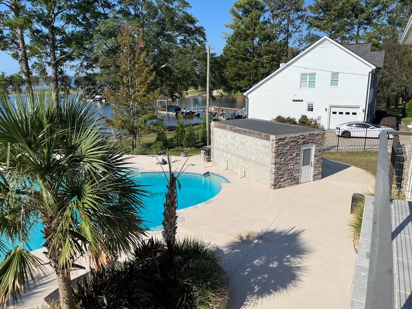 Elevated view of a curved swimming pool and broad deck, a stacked-stone pump house, palms in the foreground and a lake with a dock behind