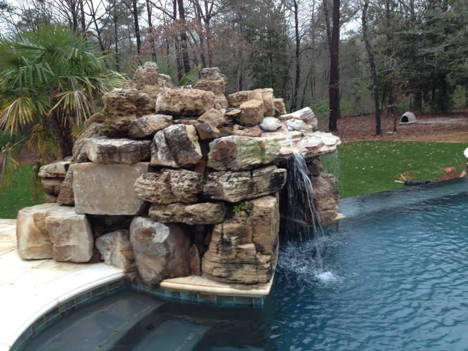 Waterfall built from stacked natural boulders spilling into the deep end of a swimming pool, with a spa in the foreground and woods behind