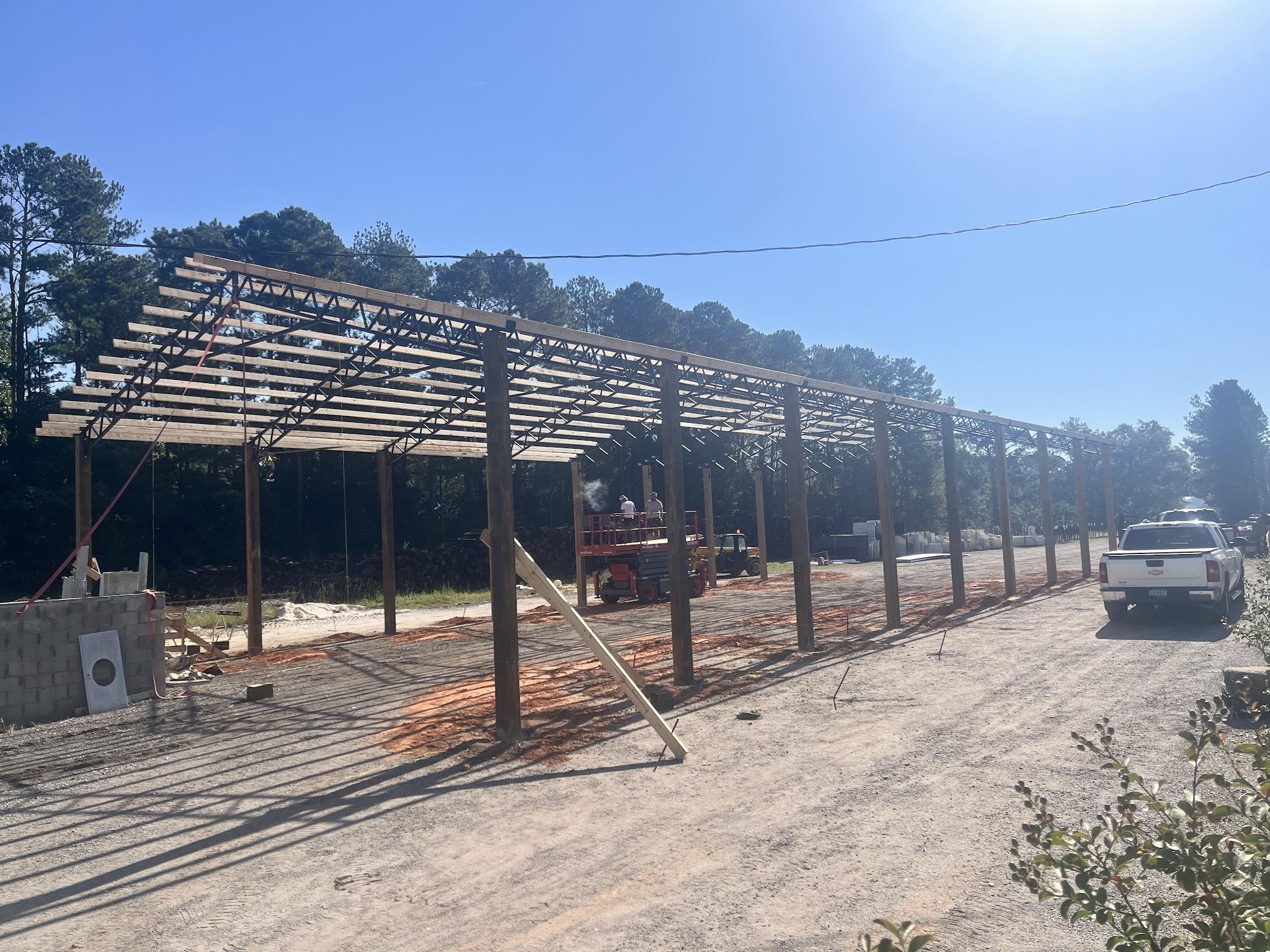 Long timber and steel frame of a covered structure under construction over a gravel yard, with a pickup truck parked at the far end and pines behind