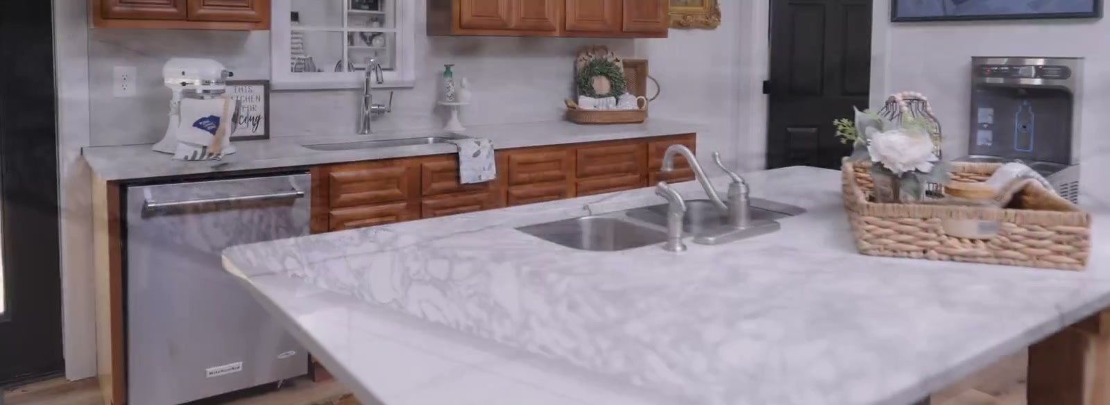 Display kitchen vignette in the showroom: a white veined stone island with an undermount double sink, wood cabinetry and a stainless dishwasher