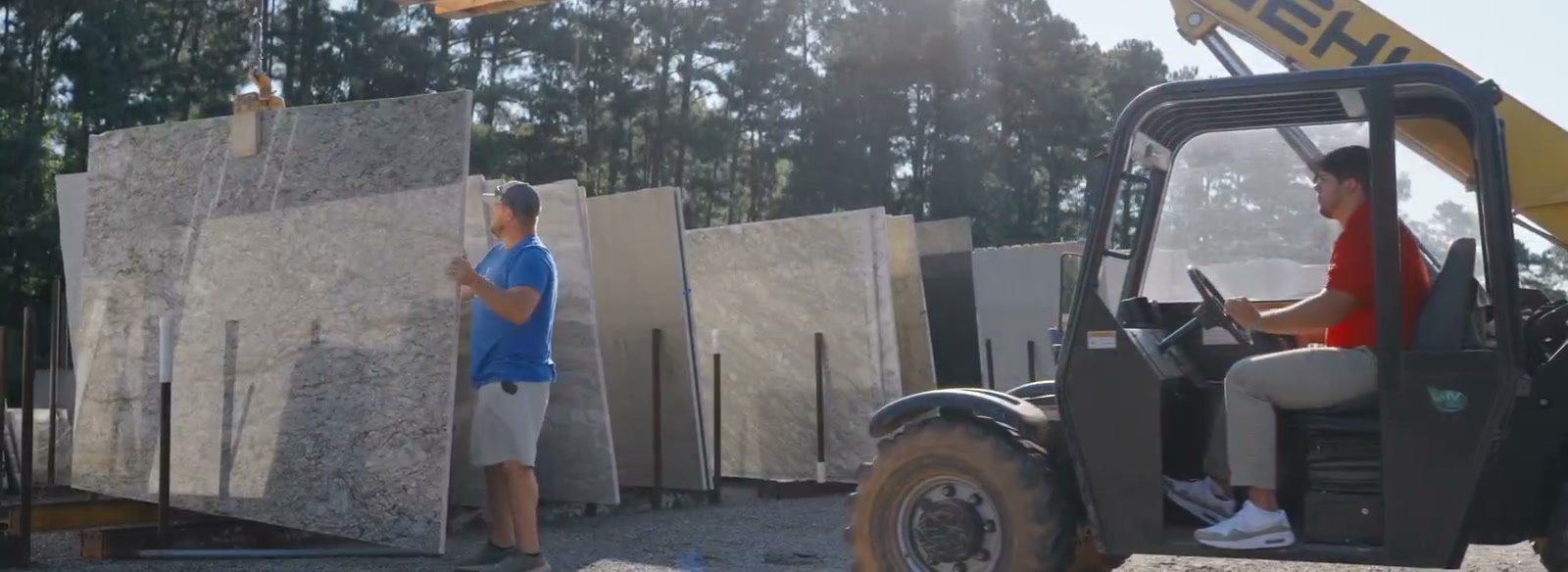 Stone yard: a man standing beside racked full-size slabs while a second man moves a slab on a forklift, pine trees behind