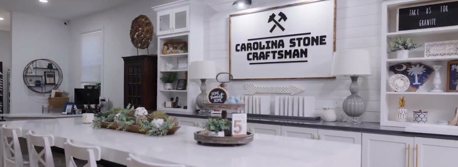 Interior of the Carolina Stone Craftsman showroom: a long white stone counter with white bar stools, built-in display shelving, and the Carolina Stone Craftsman sign mounted on the shiplap back wall