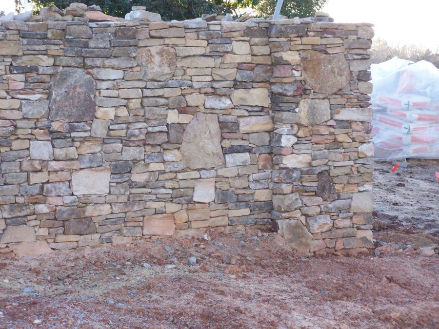 Dry-stacked fieldstone wall under construction with squared pillar sections, tan and grey stone against bare ground